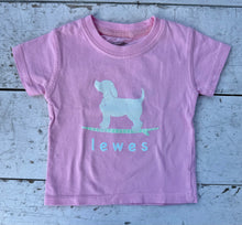 Load image into Gallery viewer, Light pink kids' short-sleeve t-shirt with a white graphic of a dog on a green and white striped surfboard, with the word 'lewes' underneath, laid flat on a rustic white wooden surface.
