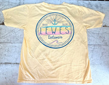 Load image into Gallery viewer, Back view of a light yellow short-sleeve t-shirt featuring a circular graphic with "Lewes Delaware Since 1631" text, an anchor, and pastel yellow, pink, and teal waves, laid flat on a rustic white wooden surface.