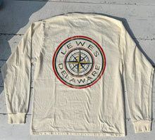 Load image into Gallery viewer, Back view of an ivory long-sleeve t-shirt featuring a large, distressed circular graphic. The graphic has a central yellow and dark blue compass rose, surrounded by 'LEWES DELAWARE' text, with red and yellow outer rings, lying flat on a distressed white wooden surface.