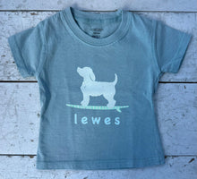 Load image into Gallery viewer, Light blue kids&#39; short-sleeve t-shirt with a white graphic of a dog on a green and white striped surfboard, with the word &#39;lewes&#39; underneath, laid flat on a rustic white wooden surface.
