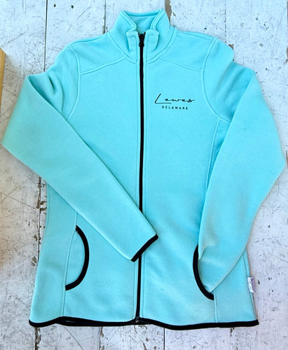 Aqua blue full-zip fleece jacket featuring 'Lewes Delaware' text, black zipper, black trim on pockets, cuffs, and hem, laid flat on a distressed white wooden surface.