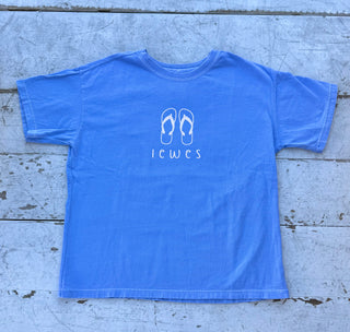 Blue short-sleeve t-shirt with a white outline graphic of two flip-flops above the word "Lewes", laid flat on a weathered white wooden surface.