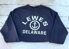 Load image into Gallery viewer, Navy blue crewneck sweatshirt featuring &#39;LEWES DELAWARE&#39; in white block letters with a white anchor graphic in the center, laid flat on a distressed white wooden surface, emphasizing coastal style and comfort.
