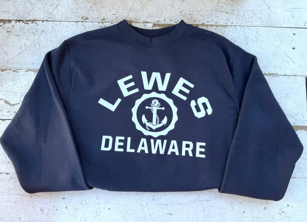 Navy blue crewneck sweatshirt featuring 'LEWES DELAWARE' in white block letters with a white anchor graphic in the center, laid flat on a distressed white wooden surface, emphasizing coastal style and comfort.