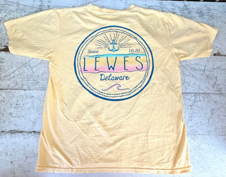 Back view of a light yellow short-sleeve t-shirt featuring a circular graphic with "Lewes Delaware Since 1631" text, an anchor, and pastel yellow, pink, and teal waves, laid flat on a rustic white wooden surface.