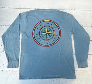 Back view of a denim long-sleeve t-shirt featuring a large, distressed circular graphic. The graphic has a central yellow and dark blue compass rose, surrounded by 'LEWES DELAWARE' text, with red and yellow outer rings, lying flat on a distressed white wooden surface.