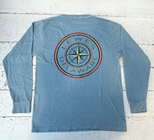 Load image into Gallery viewer, Back view of a denim long-sleeve t-shirt featuring a large, distressed circular graphic. The graphic has a central yellow and dark blue compass rose, surrounded by &#39;LEWES DELAWARE&#39; text, with red and yellow outer rings, lying flat on a distressed white wooden surface.

