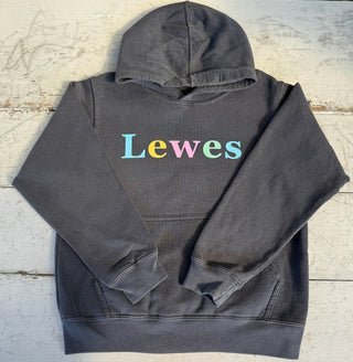 Front view of a charcoal grey unisex hoodie with a hood and a front kangaroo pocket, displaying the word 'Lewes' in multi-colored serif letters: blue L, yellow e, pink w, light green e, and light blue s, laid flat on a light wood surface.