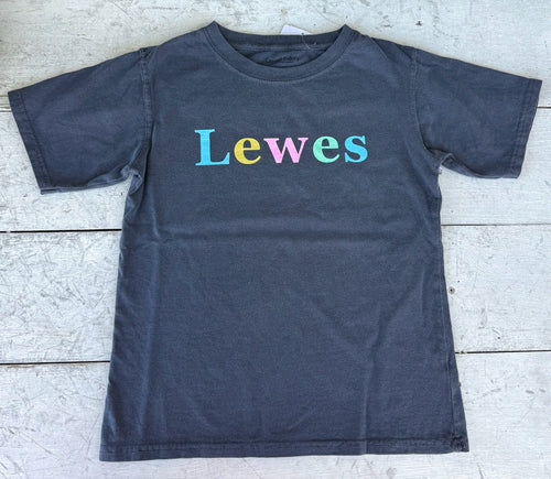 Dark gray short-sleeve t-shirt by Casual Colors, featuring 'Lewes' in pastel blue, yellow, pink, and mint green text, lying flat on white distressed wood.