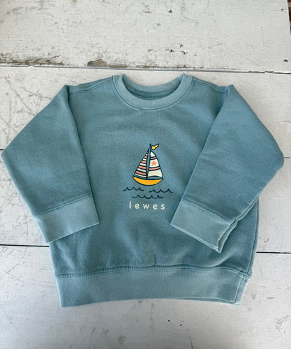 Teal blue toddler crewneck sweatshirt with a colorful sailboat and 'Lewes' graphic, displayed flat on a white distressed wooden surface.