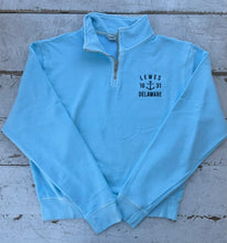 Load image into Gallery viewer, Bright light blue quarter-zip sweatshirt with white &#39;LEWES 16 ⚓ 31 DELAWARE&#39; anchor embroidery on the chest, laid flat on a rustic white wooden background.
