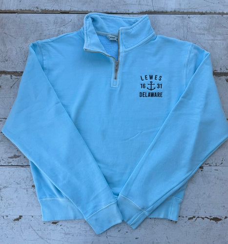 Bright light blue quarter-zip sweatshirt with white 'LEWES 16 ⚓ 31 DELAWARE' anchor embroidery on the chest, laid flat on a rustic white wooden background.