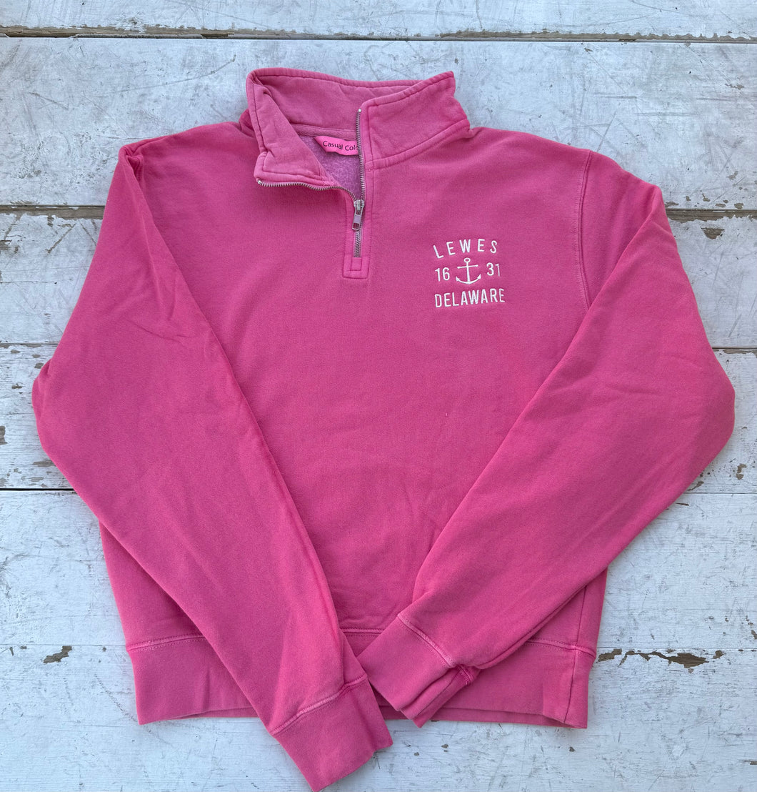 Bright hot pink quarter-zip sweatshirt with white 'LEWES 16 ⚓ 31 DELAWARE' anchor embroidery on the chest, laid flat on a rustic white wooden background.